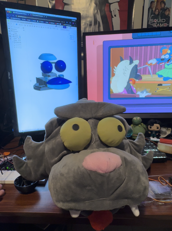 The plush animatronic head on a desk beside CAD of its eye mechanism shown on a monitor