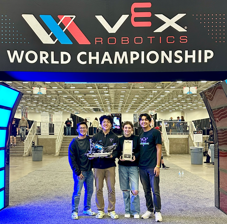 Team 5956F standing under the VEX World Championship sign, holding the robot and a trophy